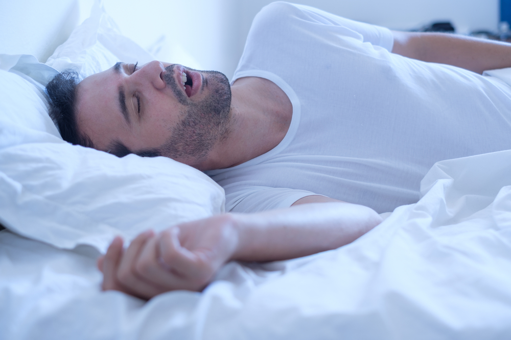 man sleeping and snoring in bed with white sheets.